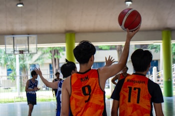 A group of young men are playing basketball indoors. Two players in orange jerseys with numbers nine and seventeen are in the foreground, with one of them holding a red basketball overhead. Other players are wearing blue jerseys, and a basketball hoop is visible in the background.