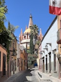 A picturesque cobblestone street flanked by colorful colonial-style buildings leads towards an ornate church with a dome and pointed spires. The Mexican flag is prominently displayed on the right, and lush greenery drapes the walls and buildings.