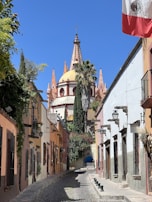A picturesque cobblestone street flanked by colorful colonial-style buildings leads towards an ornate church with a dome and pointed spires. The Mexican flag is prominently displayed on the right, and lush greenery drapes the walls and buildings.