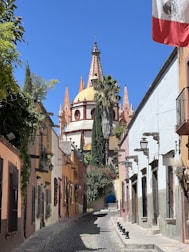 A picturesque cobblestone street flanked by colorful colonial-style buildings leads towards an ornate church with a dome and pointed spires. The Mexican flag is prominently displayed on the right, and lush greenery drapes the walls and buildings.