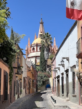 A picturesque cobblestone street flanked by colorful colonial-style buildings leads towards an ornate church with a dome and pointed spires. The Mexican flag is prominently displayed on the right, and lush greenery drapes the walls and buildings.