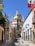 A picturesque cobblestone street flanked by colorful colonial-style buildings leads towards an ornate church with a dome and pointed spires. The Mexican flag is prominently displayed on the right, and lush greenery drapes the walls and buildings.