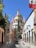 A picturesque cobblestone street flanked by colorful colonial-style buildings leads towards an ornate church with a dome and pointed spires. The Mexican flag is prominently displayed on the right, and lush greenery drapes the walls and buildings.