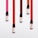 Five small bottles with black caps are arranged on a white background. Each bottle has ink spilling downward in a vibrant stripe of color varying from pink to red tones.