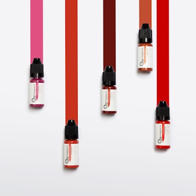 Five small bottles with black caps are arranged on a white background. Each bottle has ink spilling downward in a vibrant stripe of color varying from pink to red tones.