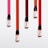 Five small bottles with black caps are arranged on a white background. Each bottle has ink spilling downward in a vibrant stripe of color varying from pink to red tones.