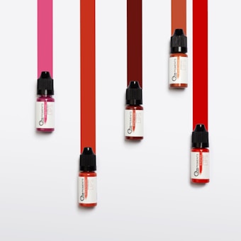 Five small bottles with black caps are arranged on a white background. Each bottle has ink spilling downward in a vibrant stripe of color varying from pink to red tones.