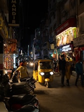 A busy urban street at night with shops lit up, displaying vibrant clothing and accessories. People walk along the street and a yellow auto-rickshaw drives through the center. The scene is bustling with activity, with various signs in different languages and parked scooters lining the sidewalk.