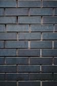 A textured wall made up of uniformly sized rectangular bricks, arranged in a horizontal pattern. The bricks are dark bluish-gray in color, with some slight variations in shading and texture. The mortar between the bricks is slightly recessed, creating a clear outline of each brick, enhancing the industrial and sturdy appearance.
