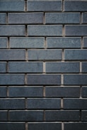 A textured wall made up of uniformly sized rectangular bricks, arranged in a horizontal pattern. The bricks are dark bluish-gray in color, with some slight variations in shading and texture. The mortar between the bricks is slightly recessed, creating a clear outline of each brick, enhancing the industrial and sturdy appearance.