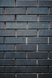 A textured wall made up of uniformly sized rectangular bricks, arranged in a horizontal pattern. The bricks are dark bluish-gray in color, with some slight variations in shading and texture. The mortar between the bricks is slightly recessed, creating a clear outline of each brick, enhancing the industrial and sturdy appearance.