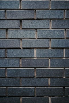 A textured wall made up of uniformly sized rectangular bricks, arranged in a horizontal pattern. The bricks are dark bluish-gray in color, with some slight variations in shading and texture. The mortar between the bricks is slightly recessed, creating a clear outline of each brick, enhancing the industrial and sturdy appearance.
