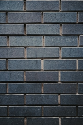 A textured wall made up of uniformly sized rectangular bricks, arranged in a horizontal pattern. The bricks are dark bluish-gray in color, with some slight variations in shading and texture. The mortar between the bricks is slightly recessed, creating a clear outline of each brick, enhancing the industrial and sturdy appearance.