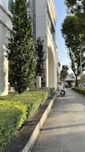 A modern building with white walls and large windows is adjacent to a neatly maintained garden with lush green shrubs and small trees. A gray sidewalk runs alongside the garden, with a motorcycle parked on it. In the background, more trees and structures are visible under a clear blue sky, suggesting a sunny day.