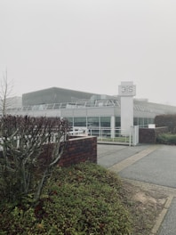 A modern building with a glass facade, surrounded by greenery and a foggy atmosphere. The structure includes a prominent sign labeled '3IS Image & Son', and the foreground features trimmed bushes and a brick wall.