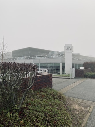A modern building with a glass facade, surrounded by greenery and a foggy atmosphere. The structure includes a prominent sign labeled '3IS Image & Son', and the foreground features trimmed bushes and a brick wall.