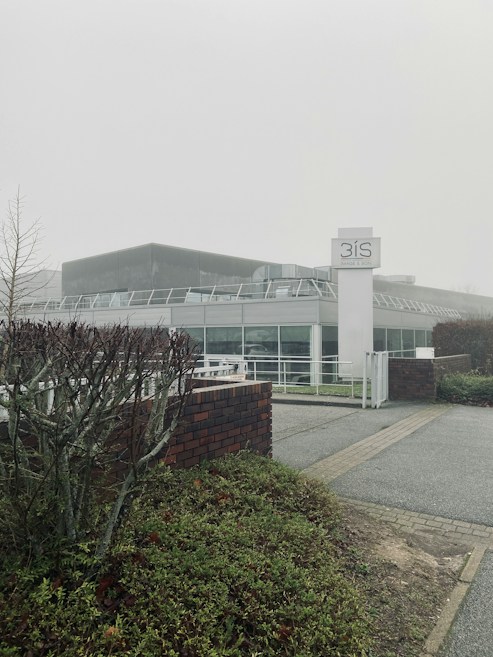 A modern building with a glass facade, surrounded by greenery and a foggy atmosphere. The structure includes a prominent sign labeled '3IS Image & Son', and the foreground features trimmed bushes and a brick wall.