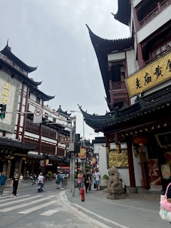 An urban street scene featuring traditional Chinese architecture with ornate, upturned eaves on the buildings. Signs with Chinese characters are visible alongside various street signs. People walk along the street, and some ride scooters. A stone lion statue is positioned near one of the buildings.