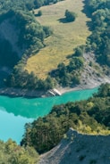 A serene landscape featuring a turquoise body of water surrounded by lush greenery and rolling hills. The trees are abundant and healthy, providing dense foliage that contrasts with the smooth water surface. Sunlight illuminates patches of grassland on the hills, creating a harmonious blend of land and water.