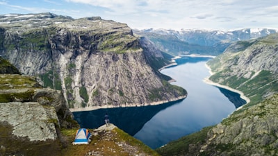 A breathtaking landscape featuring a tent pitched on a grassy cliff with a person standing near the edge, overlooking a vast fjord surrounded by steep, rugged mountains. The scene captures the serene beauty of nature with clear skies above.