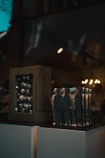 A collection of abstract sculptures displayed on a pedestal inside a dimly lit room. The sculptures are shaped like human figures made of a metallic material, arranged closely together. On the left, there is another sculpture consisting of a rectangular frame with similar metallic elements inside. The background features warm lighting fixtures that add a soft glow to the scene.