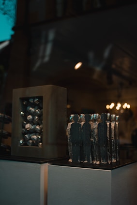 A collection of abstract sculptures displayed on a pedestal inside a dimly lit room. The sculptures are shaped like human figures made of a metallic material, arranged closely together. On the left, there is another sculpture consisting of a rectangular frame with similar metallic elements inside. The background features warm lighting fixtures that add a soft glow to the scene.