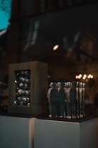 A collection of abstract sculptures displayed on a pedestal inside a dimly lit room. The sculptures are shaped like human figures made of a metallic material, arranged closely together. On the left, there is another sculpture consisting of a rectangular frame with similar metallic elements inside. The background features warm lighting fixtures that add a soft glow to the scene.