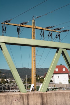 A large metal structure with electrical wiring is prominently featured, set against a backdrop of distant hills and blue sky. Below the structure, a residential neighborhood with a white house and a red roof is visible, along with trees and other buildings.