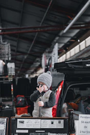 A young child wearing a gray beanie and a vest holds a small microphone while standing next to an open car trunk, surrounded by market stalls displaying various items including pendants.