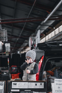 A young child wearing a gray beanie and a vest holds a small microphone while standing next to an open car trunk, surrounded by market stalls displaying various items including pendants.
