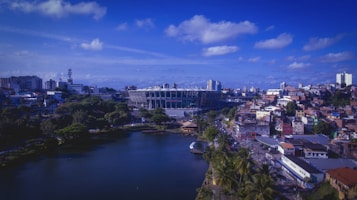 A vibrant cityscape with a large sports stadium at the center. The area around the stadium features a mix of modern and traditional buildings, with dense foliage and a peaceful body of water in the foreground. The sky is clear with scattered clouds, giving a bright and lively atmosphere.