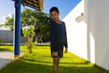 A child stands on a green lawn with a relaxed posture, holding a blue jump rope. The child is wearing a dark shirt and shorts. A blue column and some trees with sunlight shining through them are in the background, and there is a white wall on the right side casting a shadow.