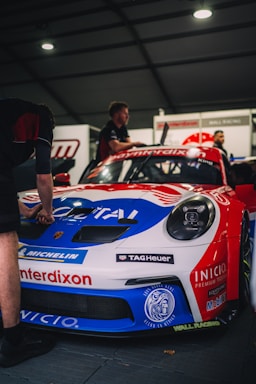 A race car in a garage, prominently featuring a Porsche with vibrant blue, red, and white sponsor logos. Two individuals are working on or inspecting the car, with a focus on the detailed branding and racing setup.