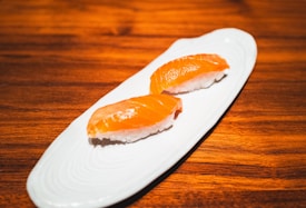 Two pieces of salmon nigiri sushi are placed on a sleek, white ceramic plate set against a warm, wooden table background. The sushi features glossy, orange salmon slices atop small mounds of white rice.