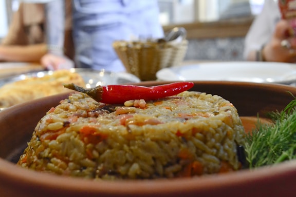 A close-up of a rice dish served in a clay bowl, garnished with a vibrant red chili pepper on top. The rice is mixed with visible ingredients like small pieces of vegetables. In the background, a blurred setting with a person and a basket filled with utensils can be seen, alongside a table setting with plates.