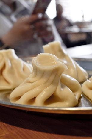 Steamed dumplings with intricate folds displayed on a metal plate. The background reveals a person holding a phone, slightly out of focus, in a dining setting.