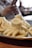 Steamed dumplings with intricate folds displayed on a metal plate. The background reveals a person holding a phone, slightly out of focus, in a dining setting.