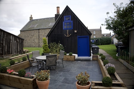 A backyard with a small wooden garden shed painted black and accented with blue features such as the door and window. The setting includes a patio area with grey tiles, outdoor furniture consisting of a round table and four chairs, and various potted plants arranged around the area. Larger stone buildings are visible in the background, and a well-maintained lawn runs alongside the patio area.