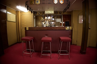 A cozy bar area with a wooden counter and three red cushioned stools. The bar is adorned with a collection of drink taps and decorative emblems hanging from the ceiling. The room has warm wooden paneling and soft lighting, creating a welcoming atmosphere. A pint of beer is visible on the counter.