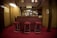 A cozy bar area with a wooden counter and three red cushioned stools. The bar is adorned with a collection of drink taps and decorative emblems hanging from the ceiling. The room has warm wooden paneling and soft lighting, creating a welcoming atmosphere. A pint of beer is visible on the counter.