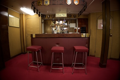 A cozy bar area with a wooden counter and three red cushioned stools. The bar is adorned with a collection of drink taps and decorative emblems hanging from the ceiling. The room has warm wooden paneling and soft lighting, creating a welcoming atmosphere. A pint of beer is visible on the counter.