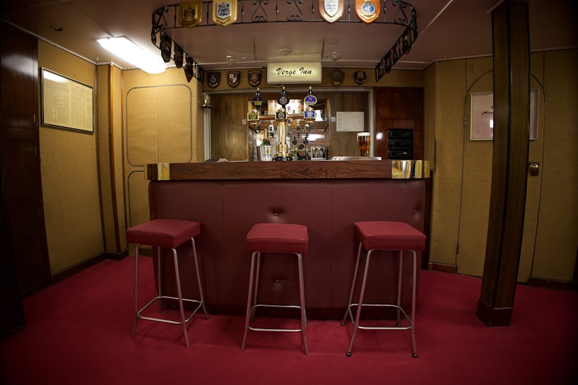 A cozy bar area with a wooden counter and three red cushioned stools. The bar is adorned with a collection of drink taps and decorative emblems hanging from the ceiling. The room has warm wooden paneling and soft lighting, creating a welcoming atmosphere. A pint of beer is visible on the counter.