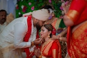 A wedding ceremony with the groom gently kissing the bride's forehead. The bride, adorned in a red and gold sari, is sitting gracefully with her hands joined. The groom is dressed in a white traditional outfit with a turban, and both wear ornate jewelry. Surrounding them are people dressed in colorful traditional attire, with a vibrant floral background.