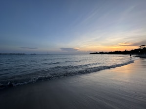Sunset over a calm Mediterranean beach with gentle waves and a small sailboat on the horizon.
