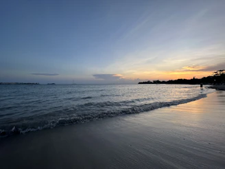 Golden sandy beach at sunset with gentle waves and a lone sailboat on the horizon.