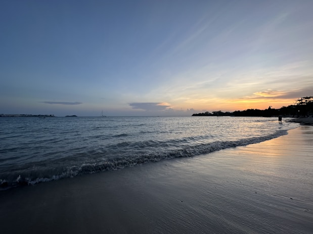 Sunset over a calm Mediterranean beach with gentle waves and a small sailboat on the horizon.