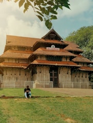 A traditional temple structure with intricate carvings and multiple tiered roofs, framed by a lush green lawn and overhanging tree branches. A person is sitting on the grass in front of the temple, suggesting a moment of relaxation or contemplation under the open sky.