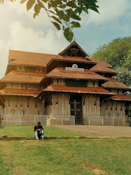 A traditional temple structure with intricate carvings and multiple tiered roofs, framed by a lush green lawn and overhanging tree branches. A person is sitting on the grass in front of the temple, suggesting a moment of relaxation or contemplation under the open sky.