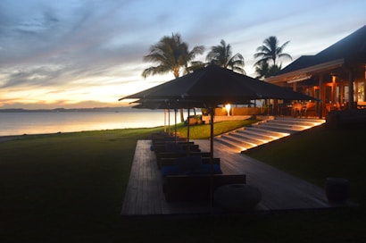 A tranquil beachside setting during sunset, featuring a wooden deck with lounge chairs and large black umbrellas. Palm trees line the background, and a modern building with warm lights is on the right. The sky displays a gradient of colors ranging from orange to blue, and the calm sea stretches across the horizon.