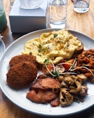 A plate of diverse breakfast items including scrambled eggs topped with greens, two crispy brown patties, roasted mushrooms, grilled bacon, and a serving of baked beans with tomatoes. The meal is set on a wooden table alongside a tissue box and a glass of water.